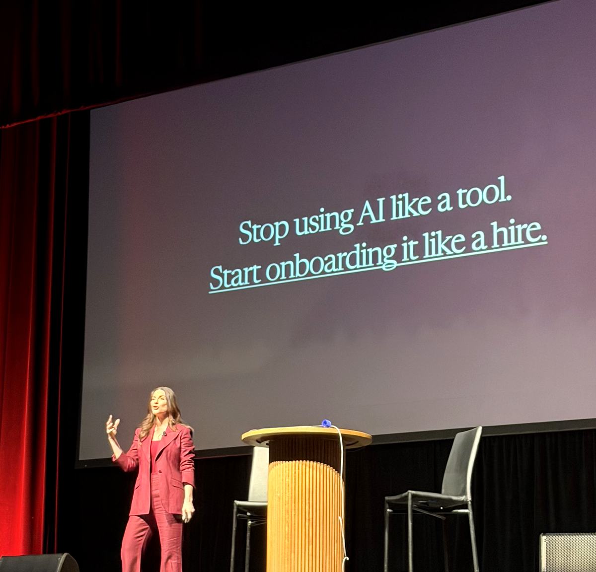 Amy Porterfield on stage with a slide reading stop using AI like a tool, start onboarding it like a hire